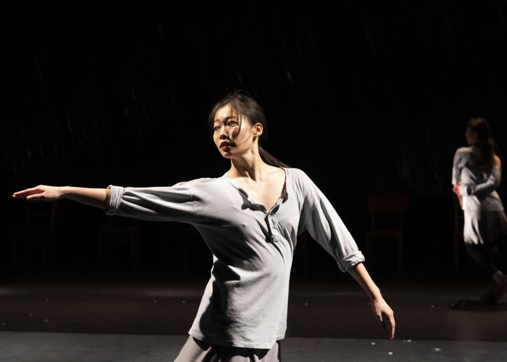 By the third section, the chairs have been lined up at the back of the stage and the choreography spreads horizontally across the space. One dancer remains seated and motionless, while the rest of the ensemble moves through fractal structures. By contrast, the fourth movement feels suddenly freer: the dancers change direction along diagonals and circles as they separate and recombine. Unison is recursively disrupted: one dancer arrives a fraction late, a phrase is altered. There is something almost euphoric about the way the choreography continually evades its own rules, just when we think we have grasped them. The music does the same, moving from a restless habanera to electronic noise and whistles. In the same spirit, a sudden blackout seems to bring the piece to a close, but then the dancers return for a brief coda that echoes the four movements.
For more than ninety minutes, the four women perform in front of us as themselves, as their own stage personas, sweating and tiring to exhaustion, far from the fictitious effortlessness often associated with virtuosity. “Rosas danst Rosas’s” strict choreographic structure never flattens the individuality of its interpreters. Its central paradox is a rigid grid inhabited by distinct personalities: impatient, vulnerable, ironic, indifferent, seductive or playful. Costumes recall rehearsal clothes or everyday dress: loose pale-blue tops, light mauve skirts, leggings, sheer socks and laced shoes. It is so Brussels that you could imagine the interpreters on the iconic Parvis de Saint-Gilles, wandering among the tables and chairs of its terraces. Low mirrors run along the sides of the stage, catching the dancers at an angle and reflecting fragments of their bodies. Within this sparse setting, emotion and meaning emerge without needing a narrative framework.