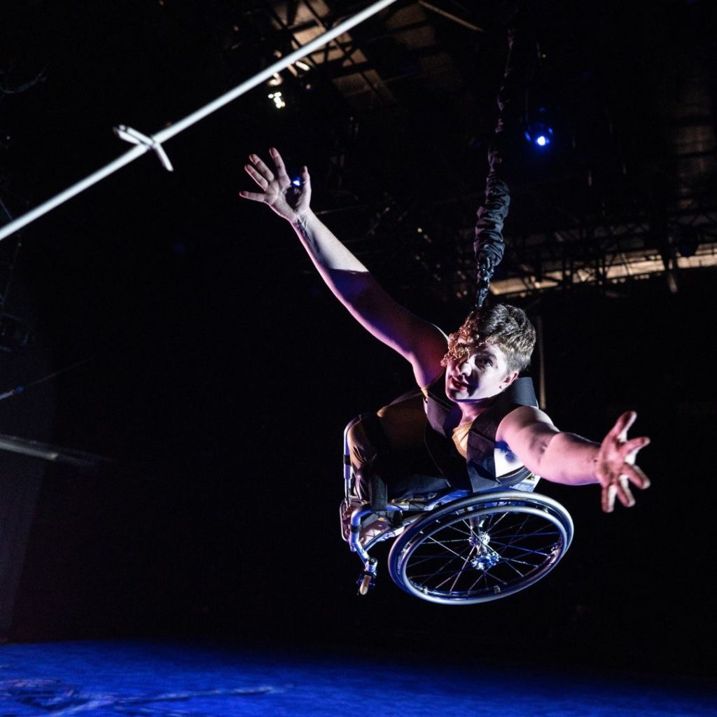 Laurel Lawson, a white dancer with very short cropped hair, bounds toward the camera: wheelchair high off the ground, arms open and reaching. A mask of delicate gold wire and lustrous pearls covers half her face. The stage floor below her is drenched in blue light; the same light gives her pale skin a light violet glow. A strand of silver barbed wire appears, close up, in the upper corner. Photo Robbie Sweeny/Kinetic Light. 